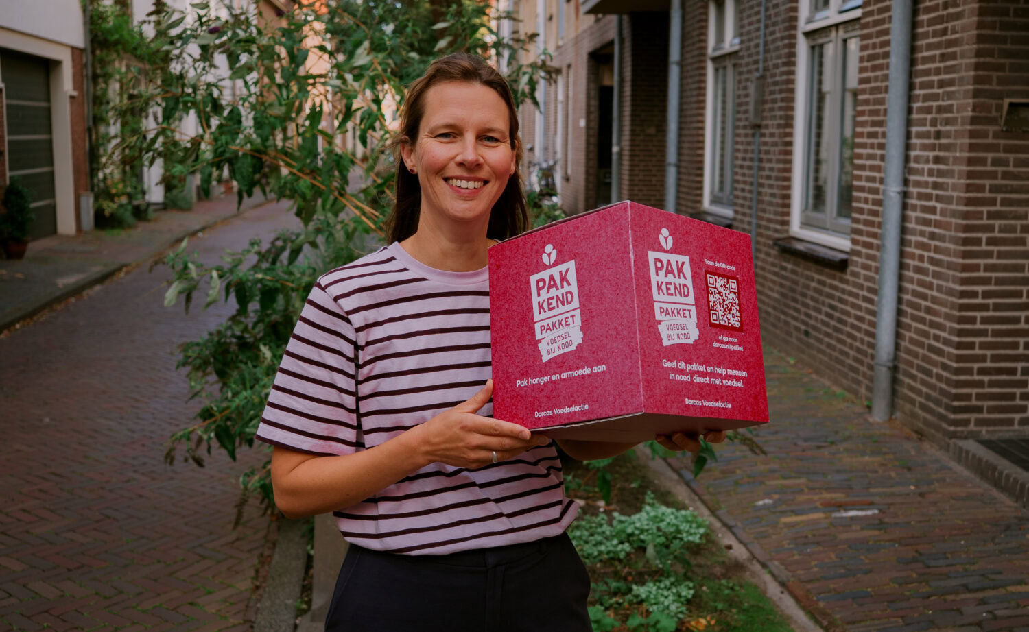 Vrouw met een glimlach houdt een roze pakket met 'Pak KEND' label, symbool voor humanitaire hulp en armoedebestrijding in Oost-Europa, passend bij Dorcas' missie.