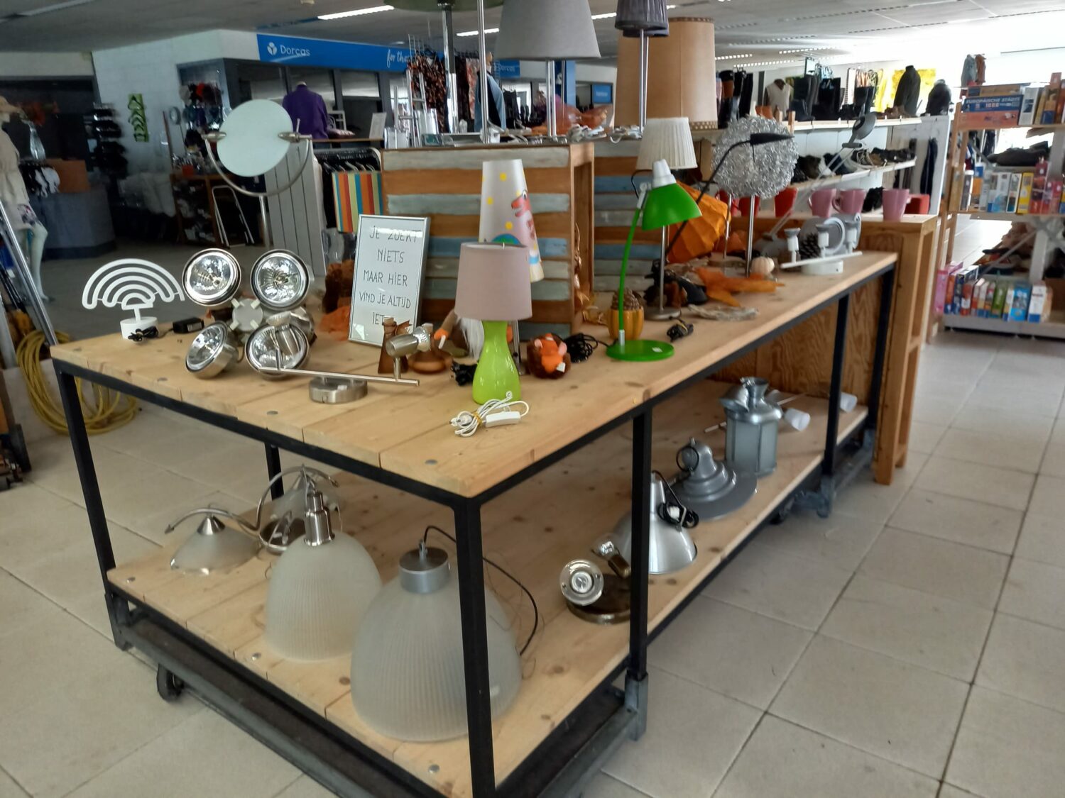 Tweedehands lampen en verlichting op een winkeltafel, geschikt voor noodhulp en armoedebestrijding, in een winkel met diverse huishoudartikelen en meubels, passend bij Dorcas' missie.