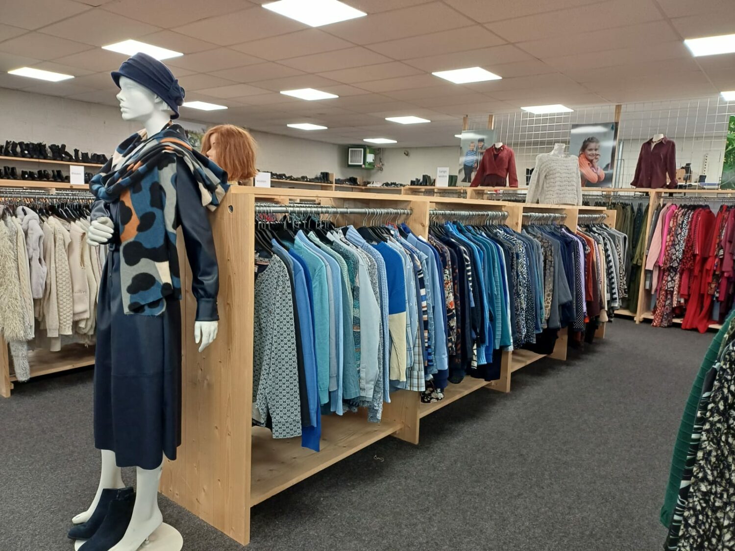 Winkel met kledingrekken vol met diverse blauwe en andere gekleurde kleding, mannequin draagt een blauwe jurk en sjaal, in een winkel voor tweedehands kleding, gericht op armoedebestrijding.