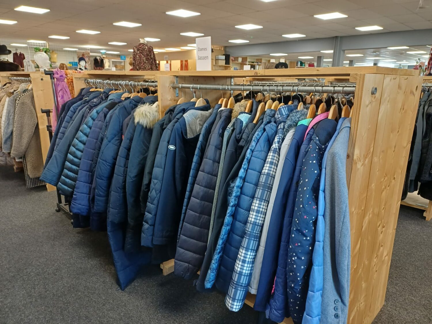 Diverse blauwe winterjassen hangen aan een houten kledingrek in een tweedehandswinkel, klaar voor verkoop. Ideaal voor mensen die behoefte hebben aan warme kleding in armoedegebieden.
