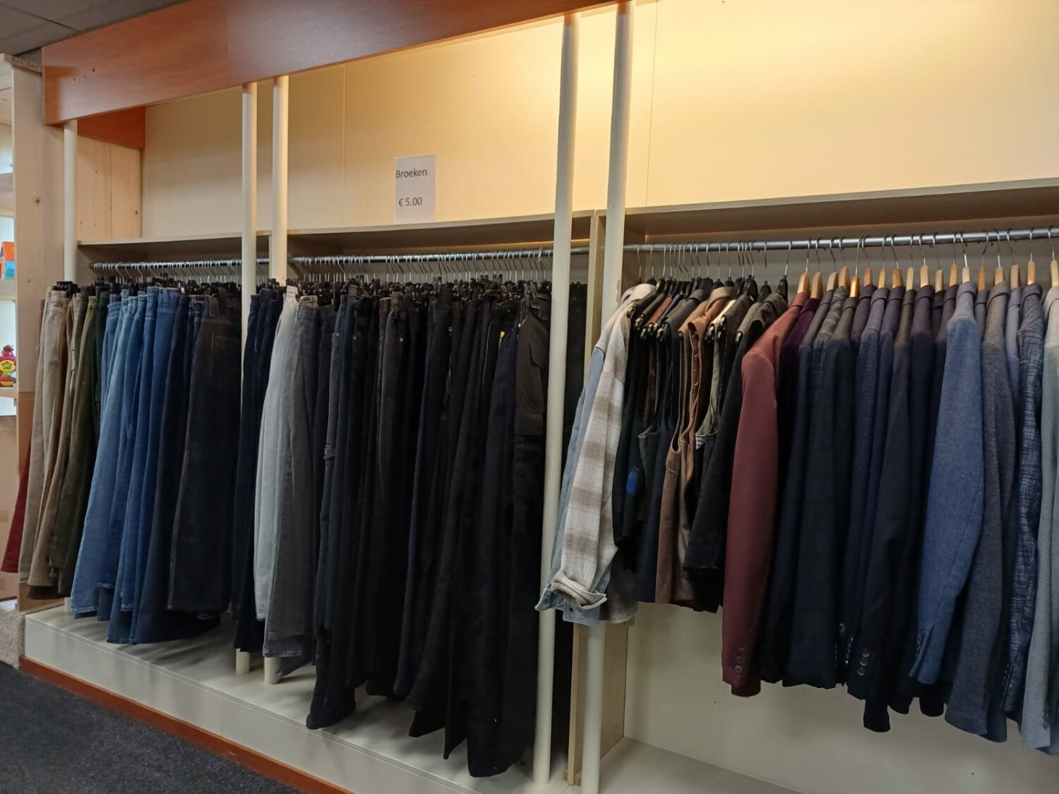 Kledingrekken met diverse jassen en broeken in een winkel, gericht op humanitaire hulp en armoedebestrijding, passend bij de missie van Dorcas.