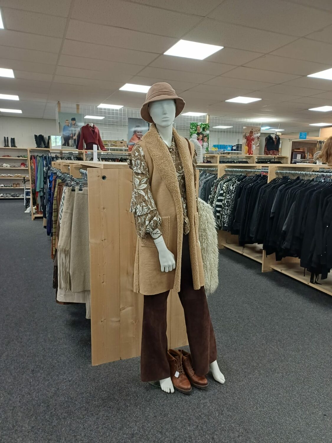 Mannequin in kledingwinkel met hoed, beige vest, blouse en bruine broek, omgeven door kledingrekken, symboliseert humanitaire hulp en armoedebestrijding.