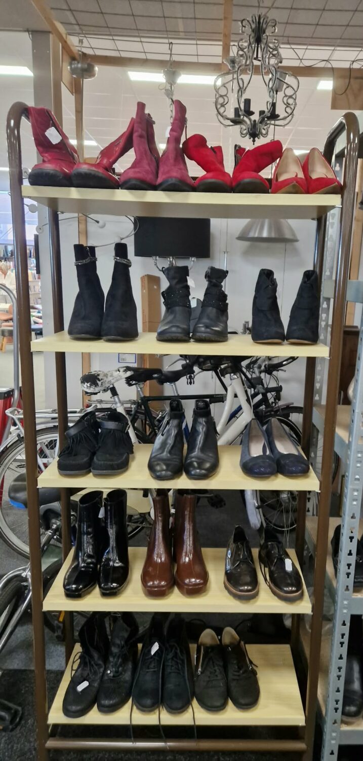 Diverse schoenen op een winkelrek, inclusief hoge hakken, laarzen en sneakers, in een winkel met een chique chandelier en grote ramen voor natuurlijk licht.