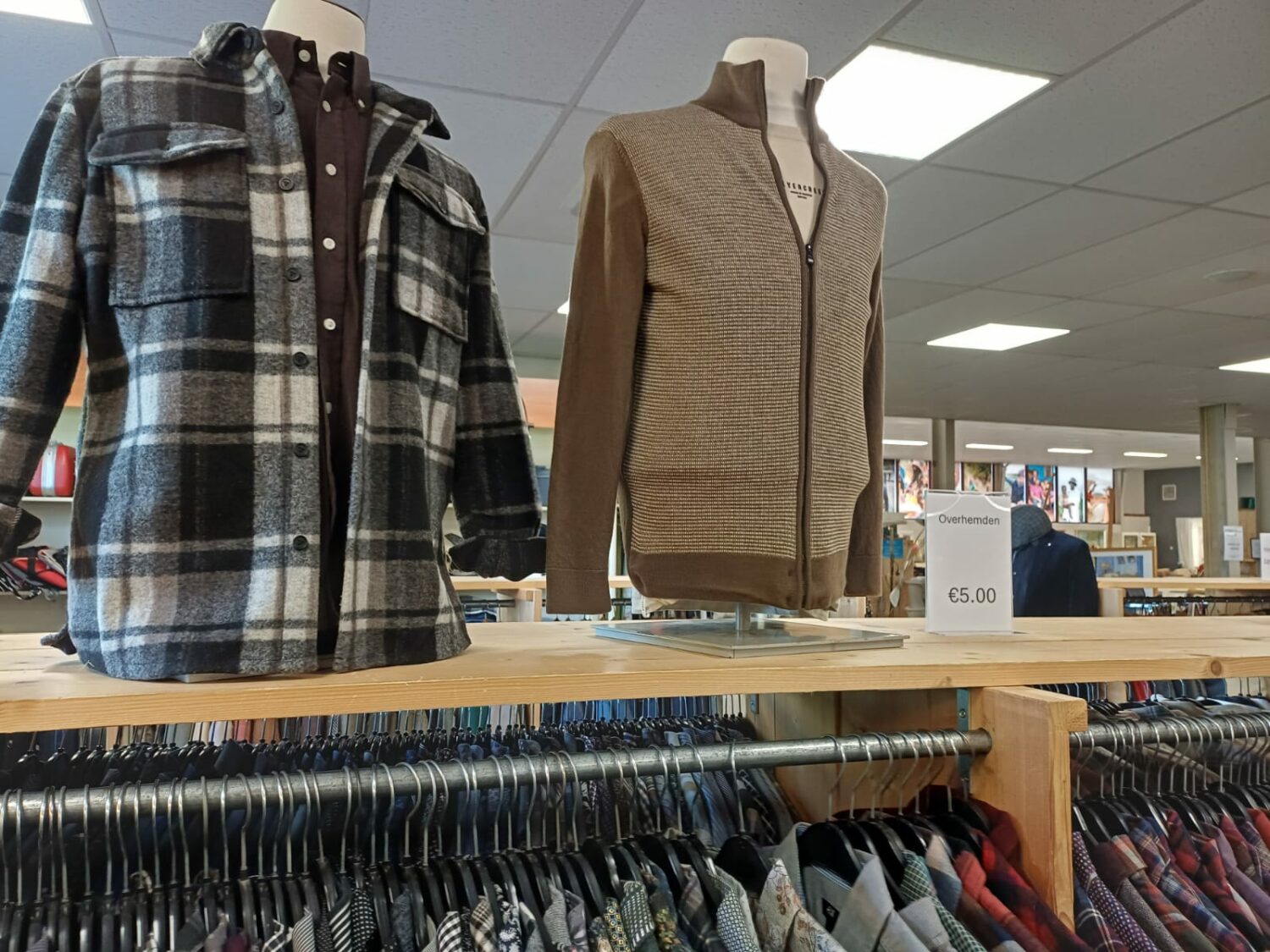 Twee herenjassen en een vest op een winkelrek, met een prijskaartje van €5,00, in een winkel die tweedehands kleding verkoopt, gericht op humanitaire hulp en armoedebestrijding.