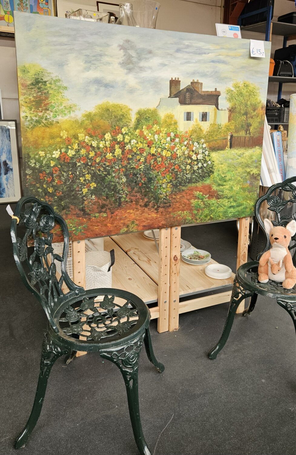 Kunstwerk van een landelijk landschap met een huis, bloemen en bomen, geplaatst op een houten standaard tussen twee zwarte tuinstoelen in een winkel of atelier, symboliseert hoop en herstel.