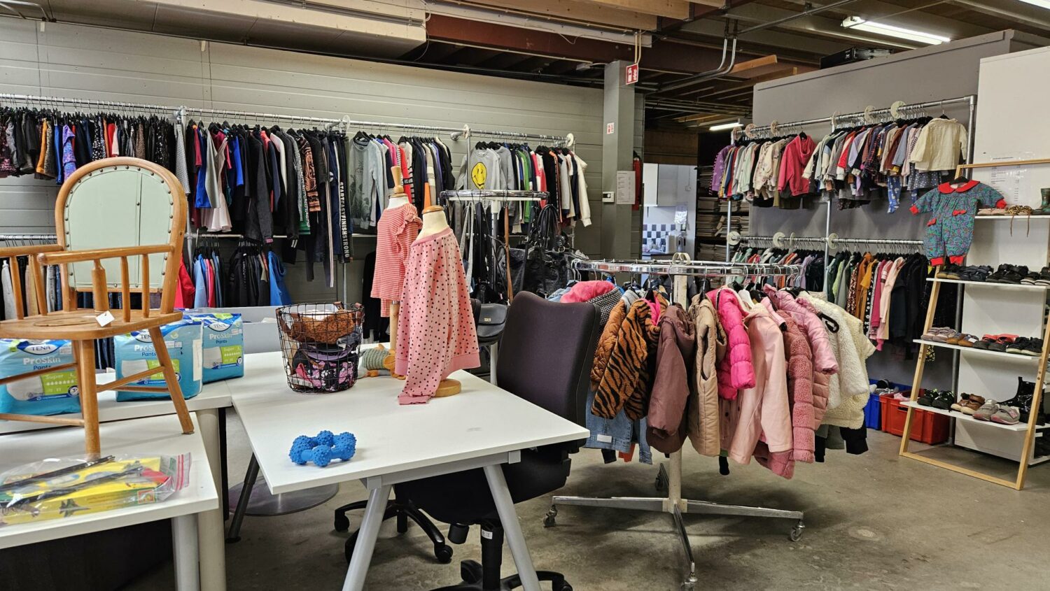In een winkel voor tweedehands kleding hangen diverse jassen en kledingstukken, met een tafel en stoelen op de voorgrond, gericht op humanitaire hulp en armoedebestrijding.