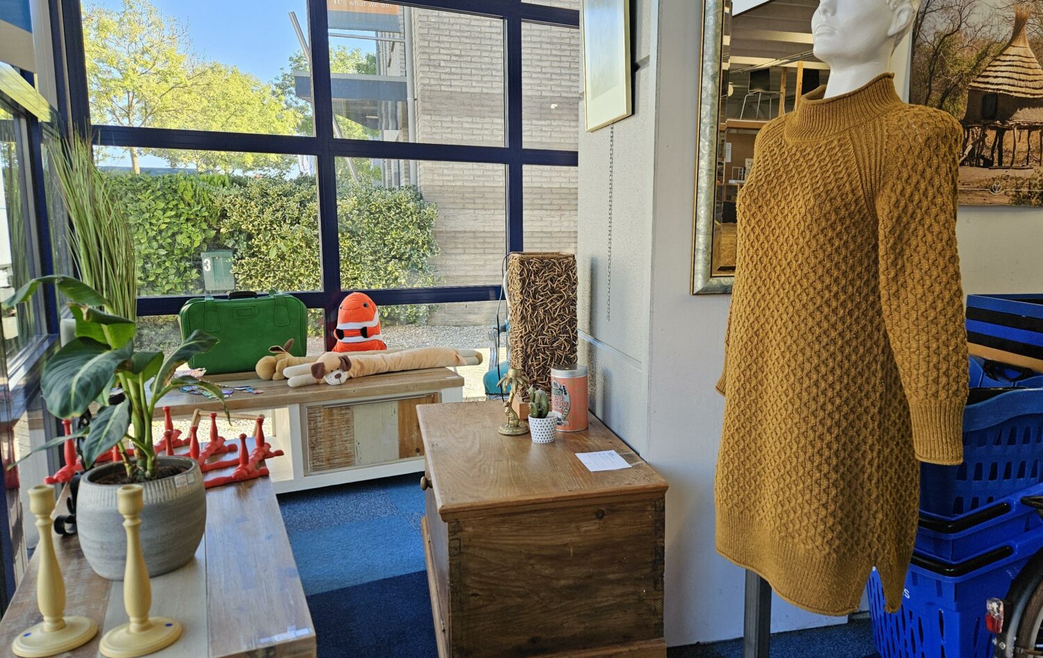 Interieur van een winkel met een gele trui op een mannequin, kleurrijke decoratie en grote ramen met uitzicht op groen, benadrukt humanitaire hulp en armoedebestrijding.