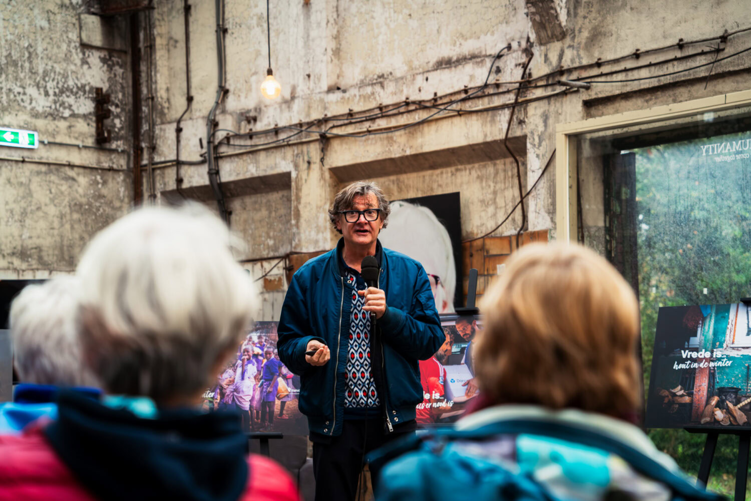 Een spreker deelt informatie over noodhulp en armoedebestrijding tijdens een Dorcas evenement, met een betrokken publiek in een industrieel ingerichte ruimte.