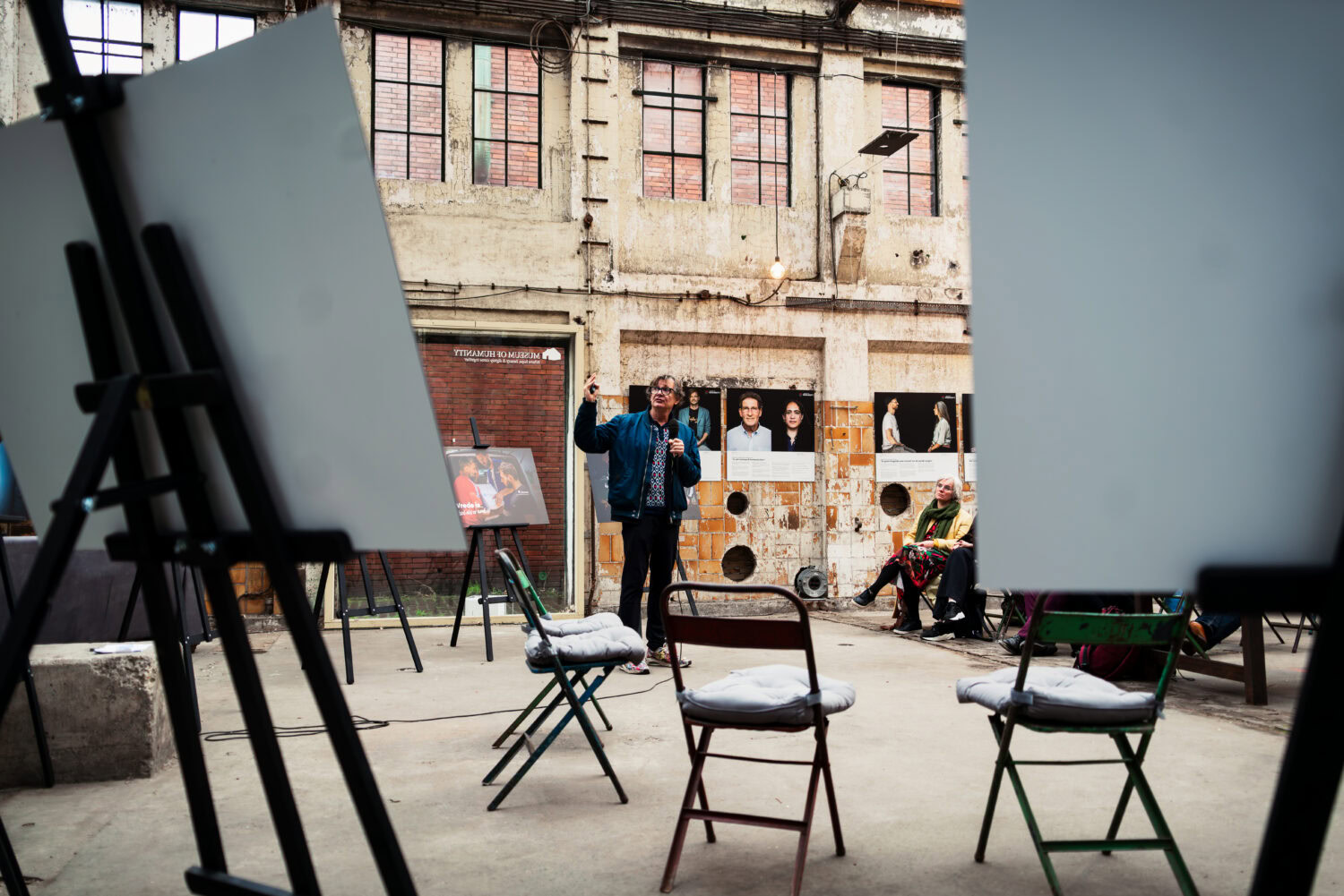 Mensen nemen deel aan een kunstworkshop in een openlucht setting, met schildersezels en stoelen, in een stedelijke omgeving die armoede en humanitaire hulp benadrukt.