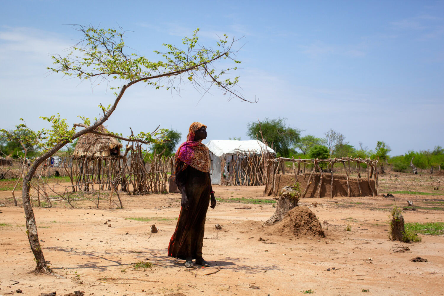 Een vrouw staat in een droog landschap met eenvoudige hutten en weinig vegetatie, illustrerend de impact van droogte en armoede in ontwikkelingslanden, passend bij de missie van Dorcas.