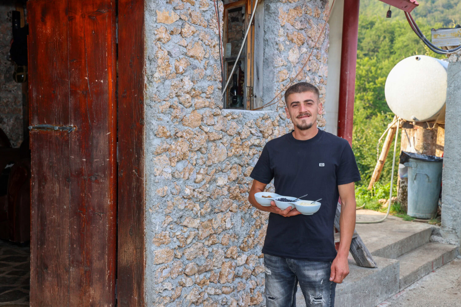 Man staat buiten een ruwe stenen muur en een houten deur, terwijl hij een schaal met eten vasthoudt. Context: Dorcasprojecten voor noodhulp en ontwikkeling in Oost-Europa.