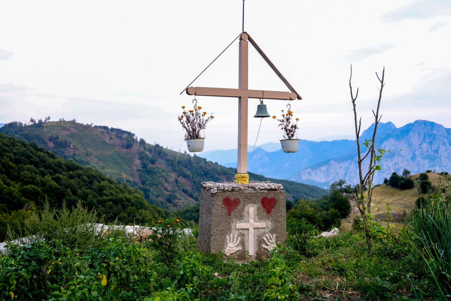Nederige kruisconstructie met bloemenbakken tegen berglandschap. Symboliek van hoop en hulpverlening in een rurale omgeving, passend bij Dorcas missie en humanitaire zorg in Oost-Europa, Oost-Afrika &.