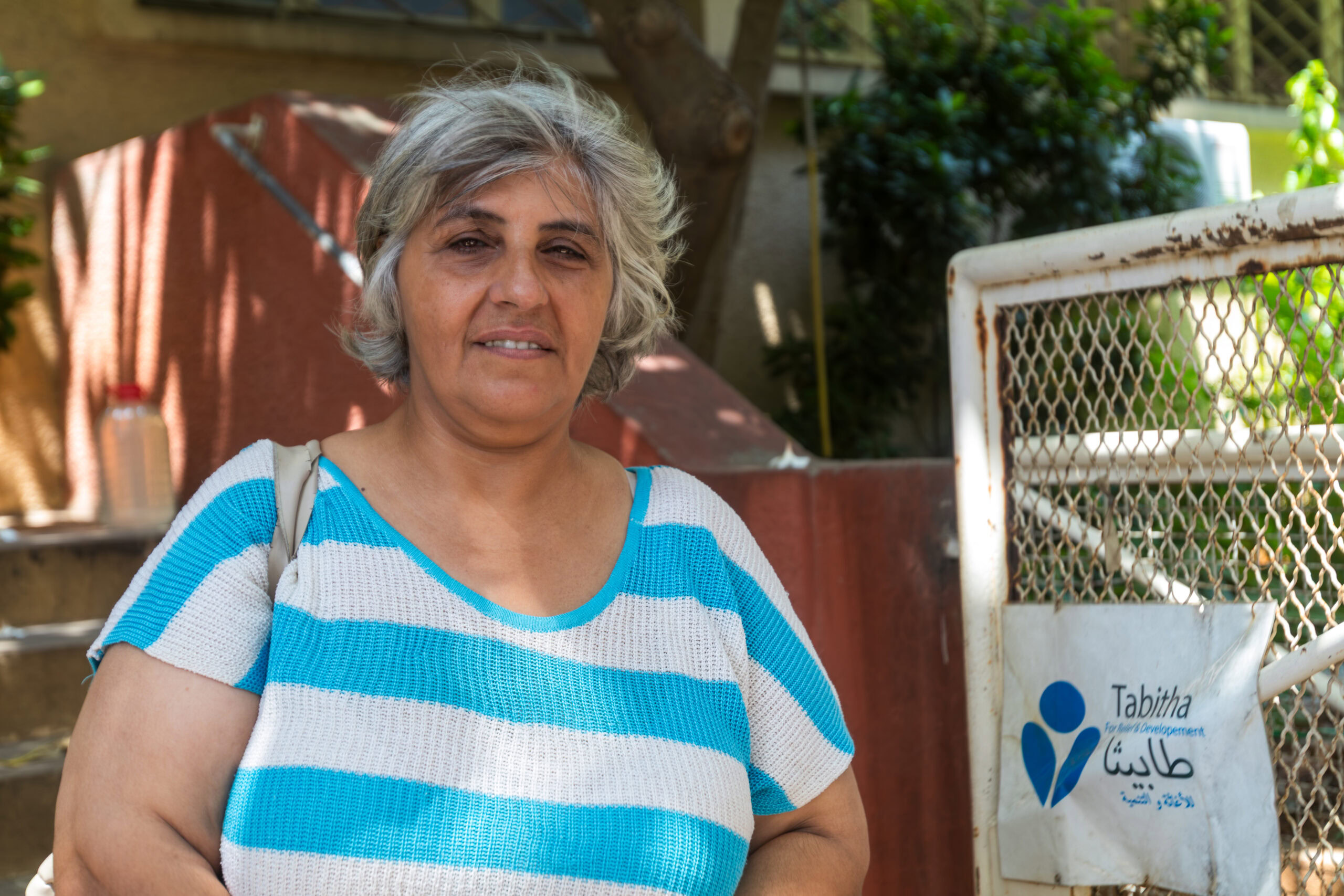 Mona uit Libanon ontvangt humanitaire hulp en steun, zichtbaar in haar vriendelijke uitdrukking en de omgeving met een hulporganisatiebord, symboliserend armoedebestrijding.