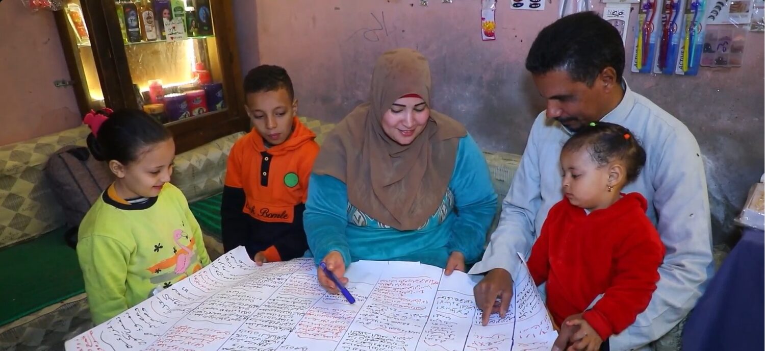Hulpverleners en dorcas-medewerkers bespreken samen met een gezin een schrift met inkomsten- en uitgavenoverzicht, terwijl kinderen meekijken in een huiselijke setting.