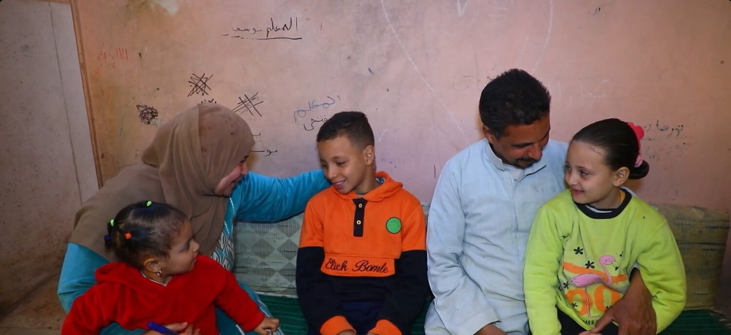 Gezinsmoment in een dorpshuis: volwassene en twee kinderen zitten op een bank, terwijl een vrouw en een man hun aandacht geven aan de kinderen en lachen; sfeer van zorg en gemeenschap in een humanitär.