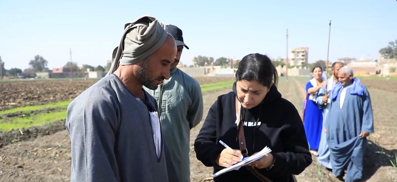 Hulpverleners van Dorcas registreren vervoer- en noodhulpacties in een veldgebied in Oost-Afrika; een vrouw schrijft notities terwijl een man naast haar de situatie bekijkt, met anderen op de tweede背景.