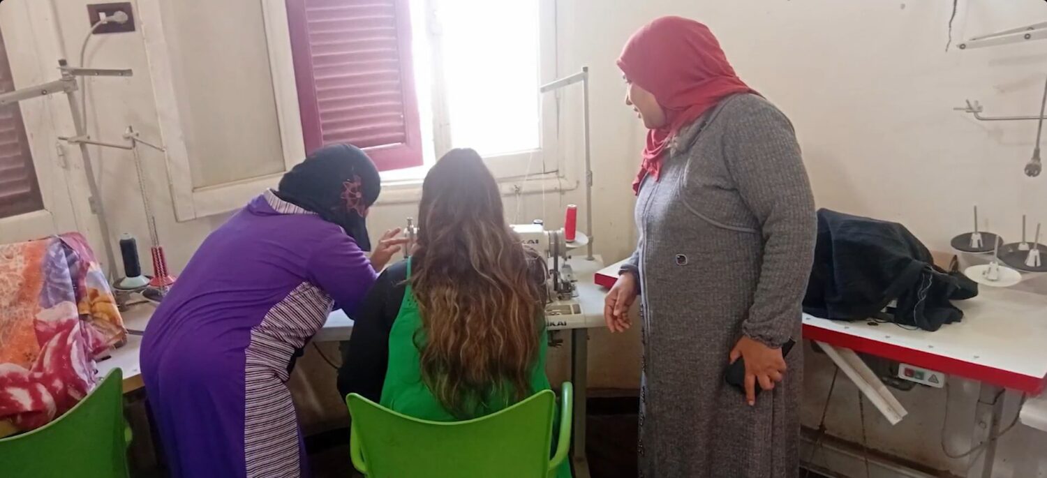 Vrouwelijke vrijwilligers helpen een student in een laboratoriumruimte met onderzoeksmateriaal; centraal figuur met rode hoofddoek observeert nabij een werktafel.