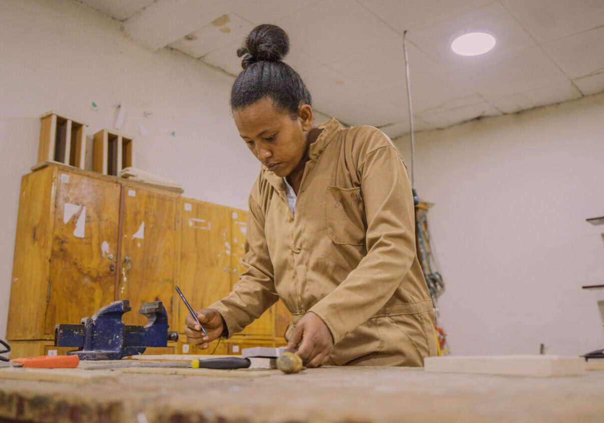 Een vrouw die geconcentreerd werkt aan een houten project in een werkplaats, met gereedschap en houtmaterialen op de tafel, onder goede verlichting.
