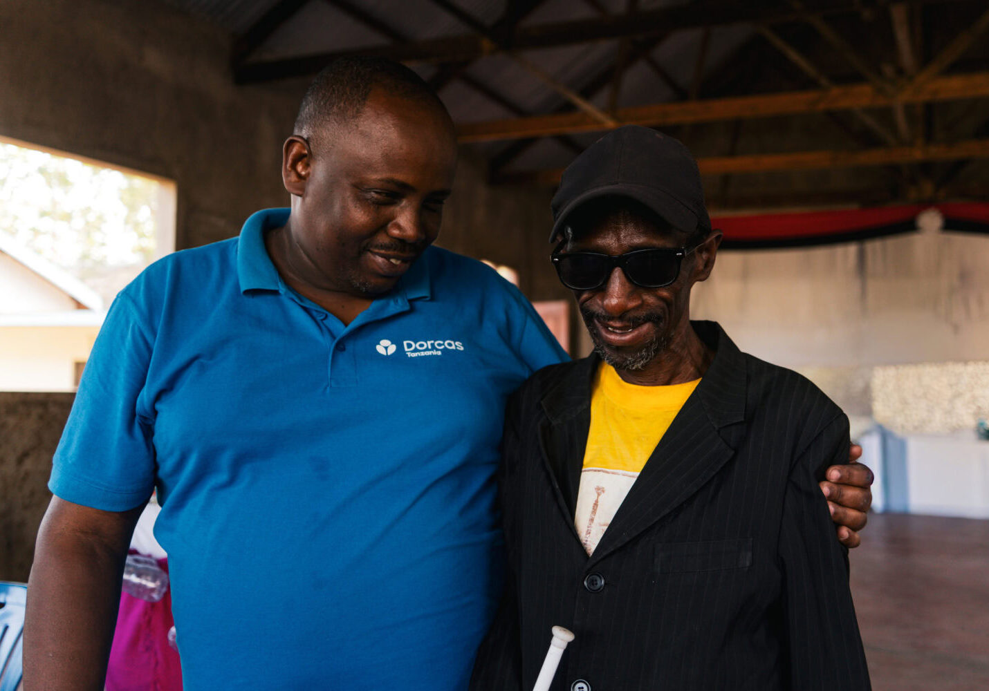 Een man in een blauwe Dorcas-polo en een man in een zwarte jas en zonnebril delen een moment van verbondenheid en samenwerking.
