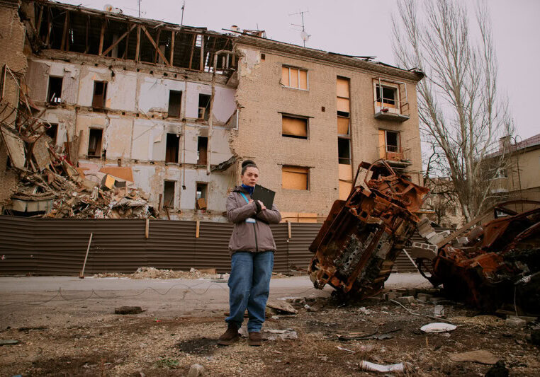 Een man staat met een tablet voor een verwoeste woning en auto, omgeven door schade en puin, illustrerend de impact van een ramp of incident op de omgeving.