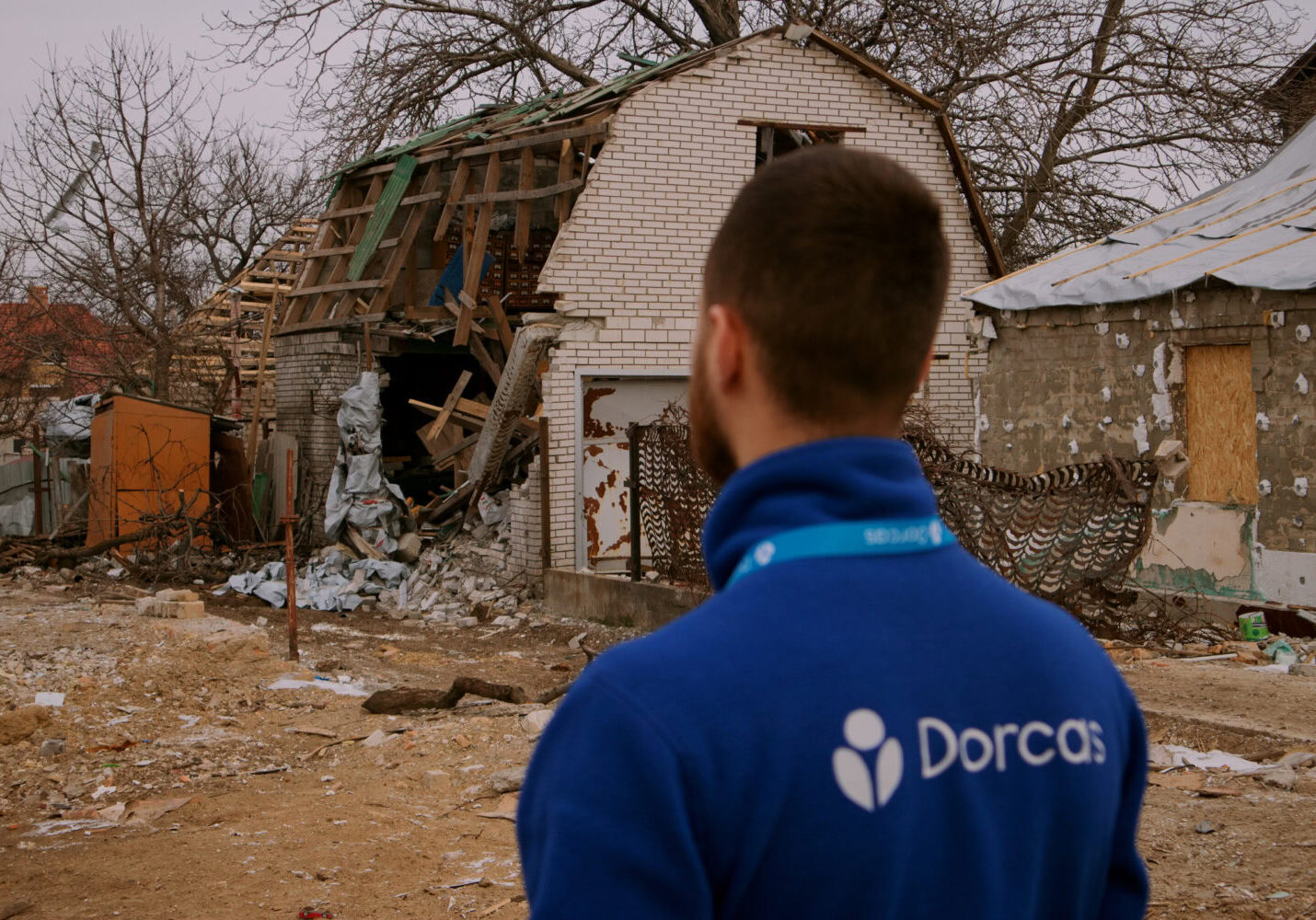Een man met een blauwe Dorcas-jas kijkt naar een verwoest huis dat beschadigd is door de oorlog in Oekraïne, symboliserend de hulp en wederopbouw door Dorcas.