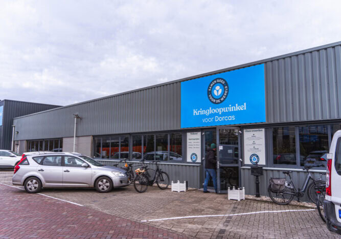 Kringloopwinkel voor Dorcas in een industrieel gebouw met parkeerplaatsen, fietsen en een blauwe bordgevel met logo en openingstijden, gericht op hergebruik en duurzaamheid.