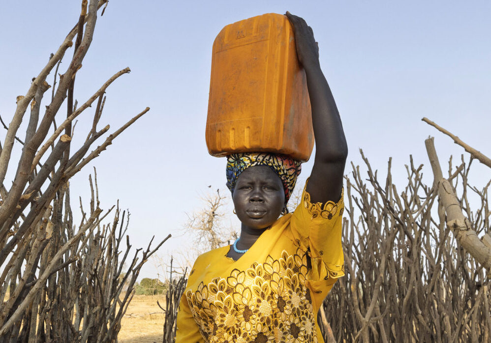 Een vrouw draagt een watercontainer op haar hoofd in een droog landschap met dorre bomen.