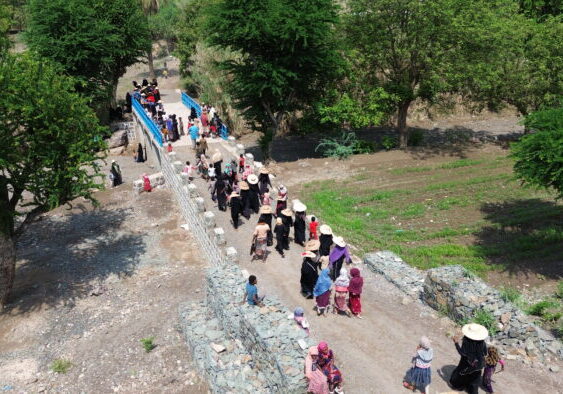 DCIM104MEDIADJI_0005.JPG Mensen lopen over een klein bruggetje door een groene omgeving met bomen en akkers.