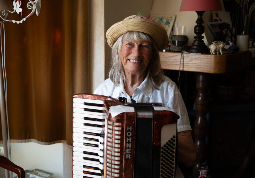Een oudere vrouw met witte haren en een strooien hoed speelt op een accordeon in een gezellige woonkamer.