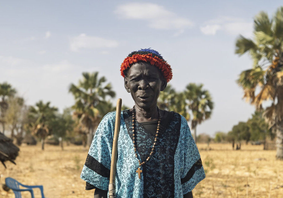Een vrouw in traditioneel kledingstuk met een bloemrijke hoofdband, omgeven door palmbomen en een dorpslandschap, symboliseert lokale cultuur en gemeenschapsontwikkeling.