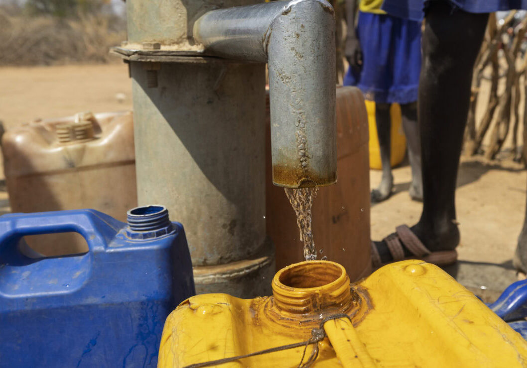 Een waterpunt met gele en blauwe jerrycans onder een waterkraan in een dorre omgeving.