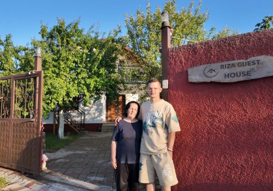 default Een foto van een vrouw en een jonge man voor de ingang van Riza Guest House, omgeven door groene bomen en een heldere blauwe lucht.