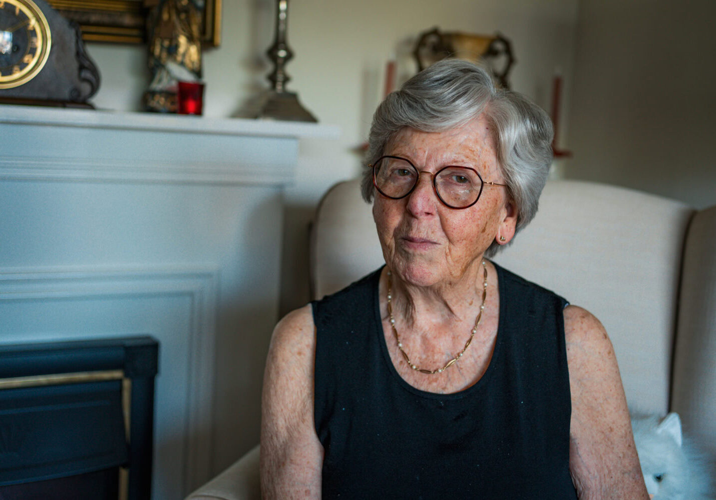 Zilverkleurige oudere vrouw met bril zit ontspannen in een woonkamer, draagt een zwart top en sieraden, sfeer van warmte en huiselijkheid.