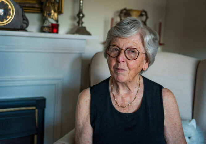 Zilverkleurige oudere vrouw met bril zit ontspannen in een woonkamer, draagt een zwart top en sieraden, sfeer van warmte en huiselijkheid.