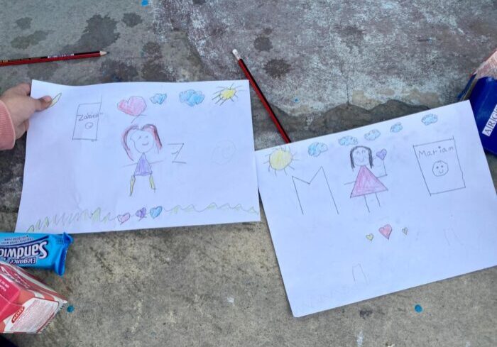 Een kindertekening met kleurrijke schetsen van mensen, zon en hartjes, gemaakt op een betonnen vloer met potloden en snacks op de achtergrond.