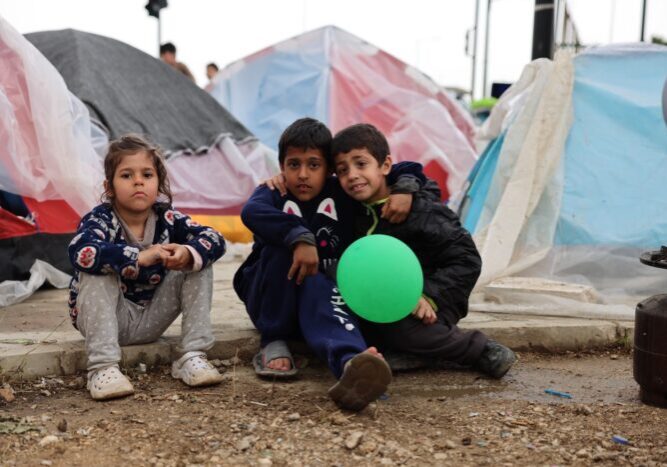 Drie kinderen zitten op de grond tussen tenten in een vluchtelingenkamp, met één kind dat een groene ballon vasthoudt, en zien er vermoeid maar hoopvol uit.