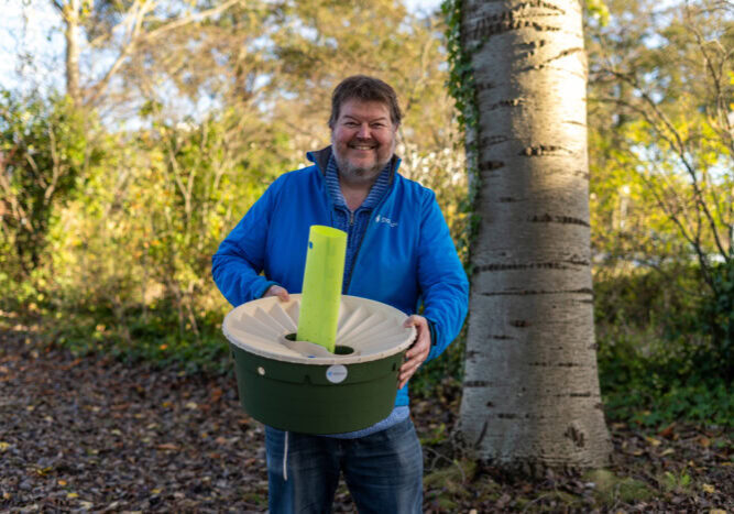 Man met een grote glimlach houdt een groene en witte container met een grote groene pen erin, buiten in een bosrijke omgeving met bomen en herfstkleuren.