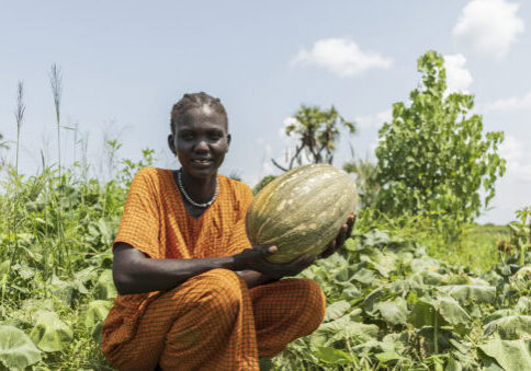 Gezicht van een vrouw in traditionele kleding die een grote watermeloen vasthoudt in een groentetuin.